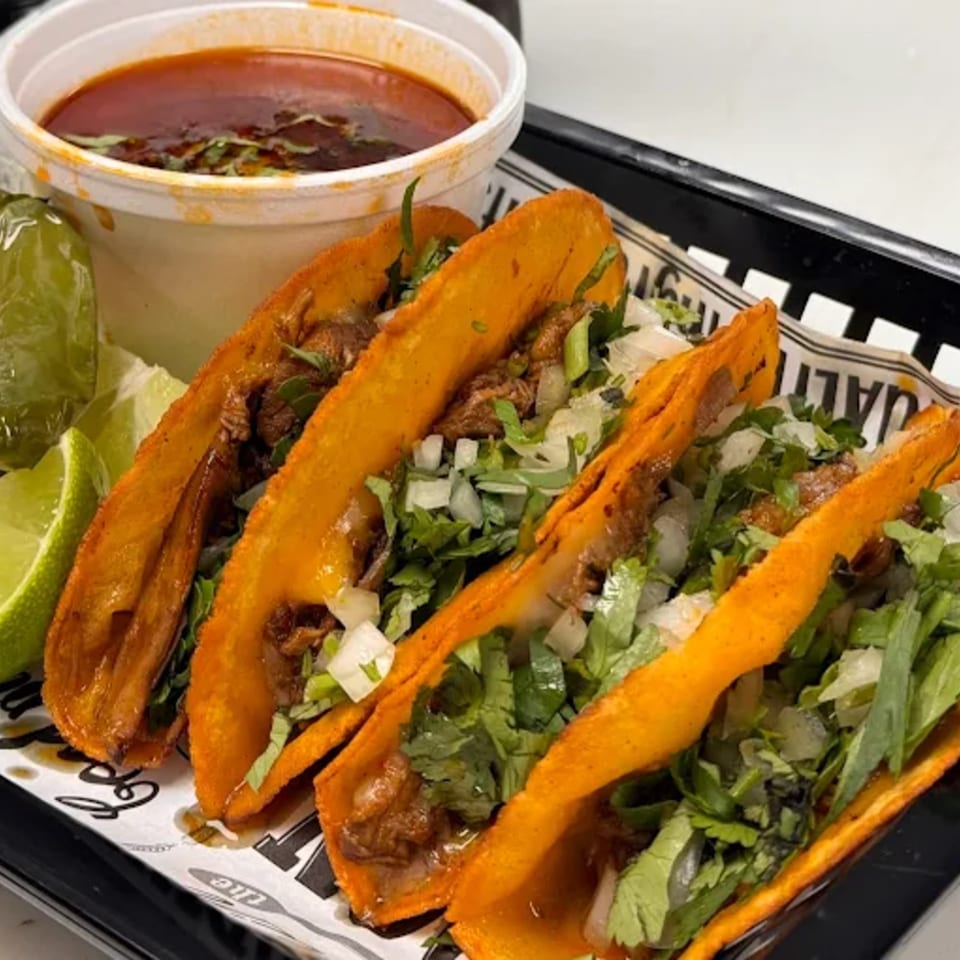 Three birria tacos with cilantro, onions, lime wedges, and a side of consommé from HP Taco Shop.