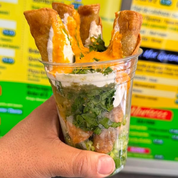 A hand holds a clear cup filled with taquitos, guacamole, and sauces from HP Taco Shop.