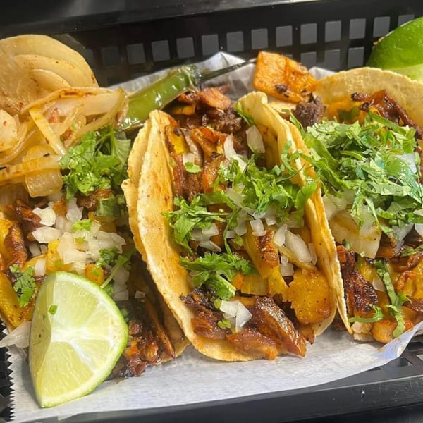 Three HP Taco Shop tacos with meat, onions, cilantro, and lime wedges, served with grilled onions and a pepper.